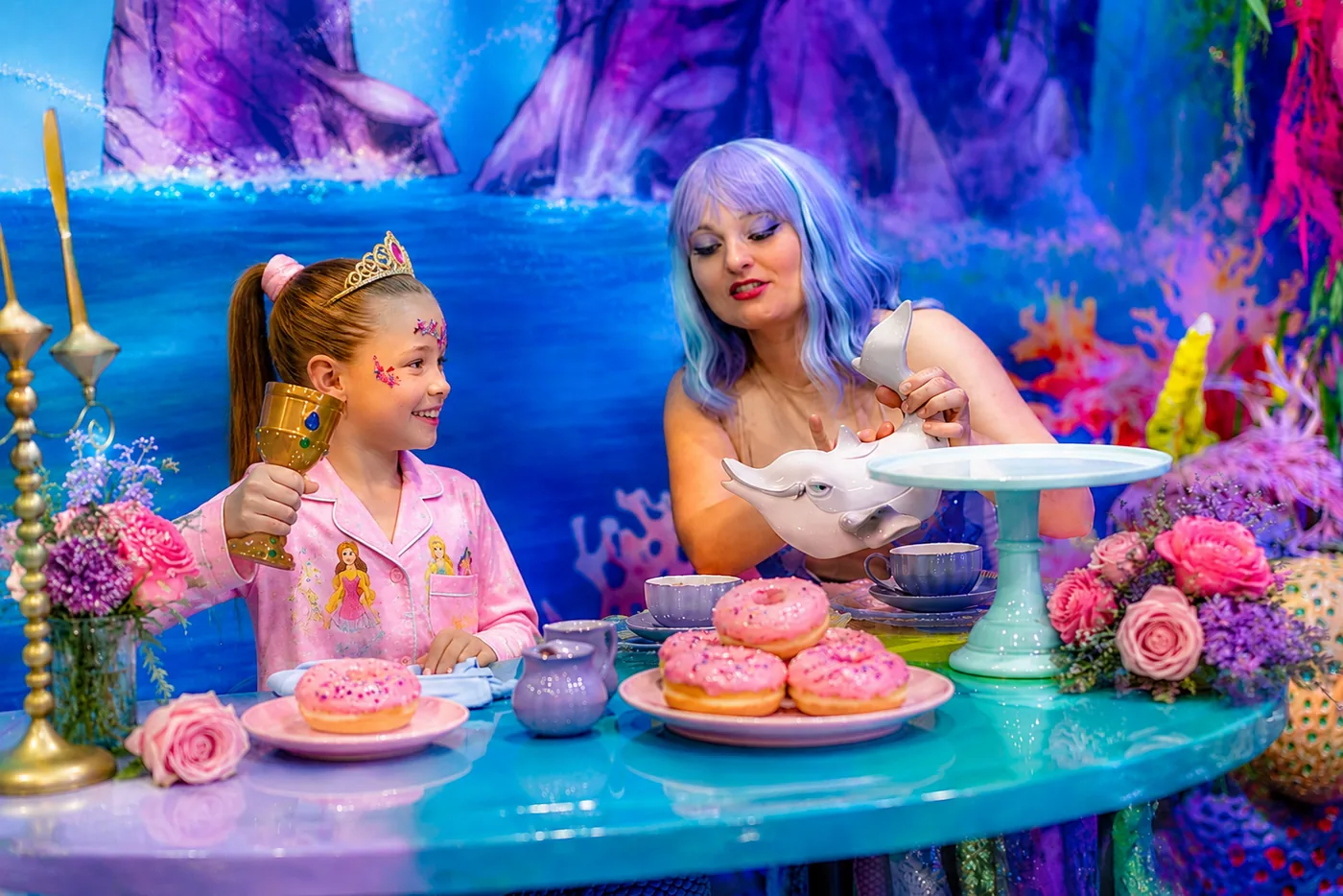 A mermaid pouring morning tea from a dolphin teapot beside pink donuts at the Mermaid Breakfast Tea Party