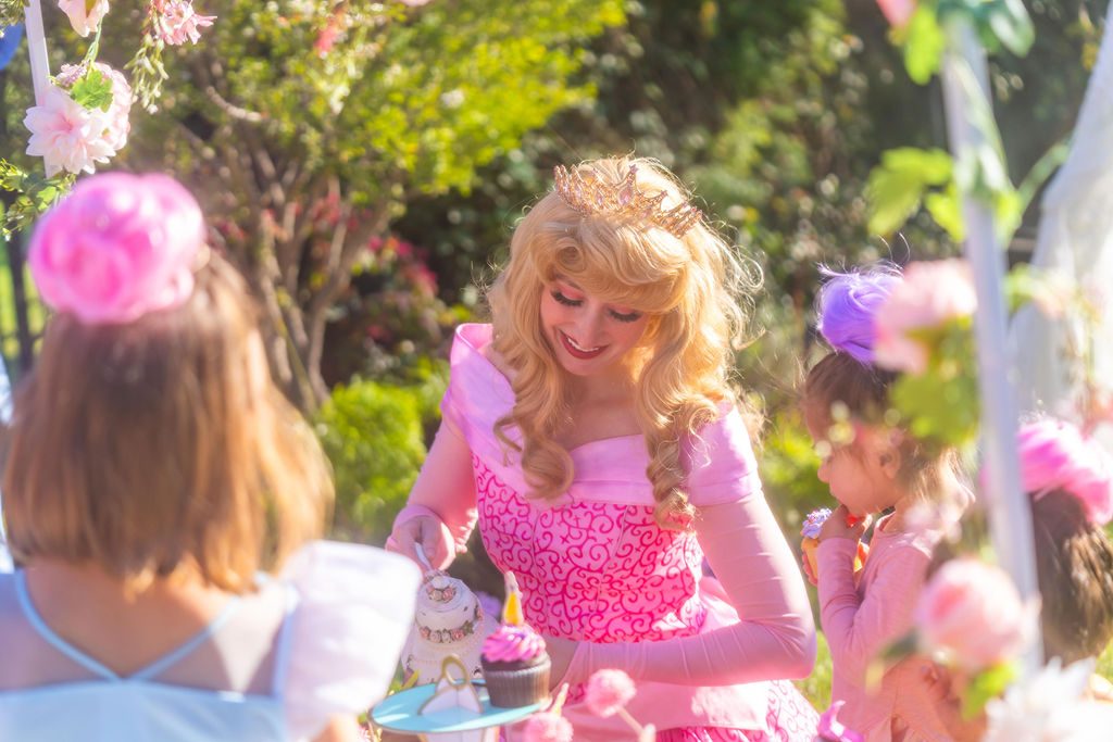 A host character kneeling beside a child at her garden birthday party