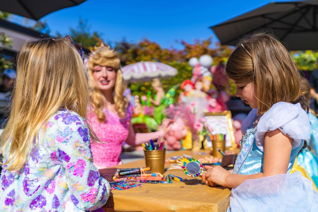 A parent watching from the side while a character helps a child at the craft table