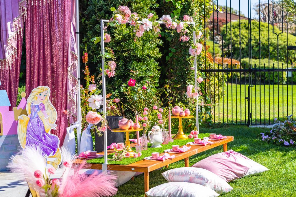 Decorated picnic tables, pillow seating and a floral backdrop built in a garden before the guests arrive