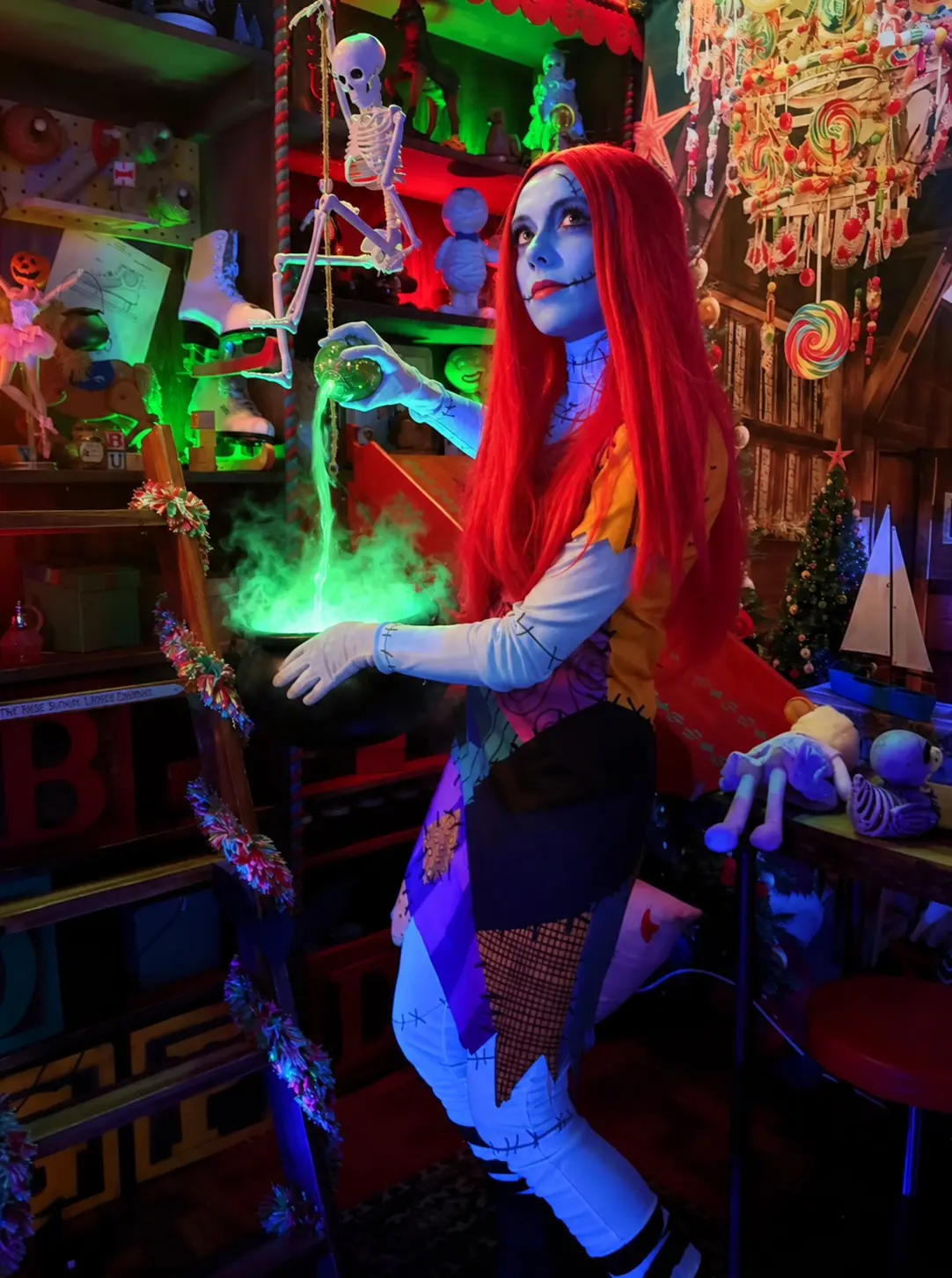 Sally, with her long red hair and patchwork dress, pours a glowing green potion into a misty cauldron in Santa&rsquo;s toy workshop, with a skeleton on a swing and giant lollipops overhead